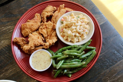 A red plate with fried chicken, macaroni salad, green beans, and a small bowl of mustard sauce.