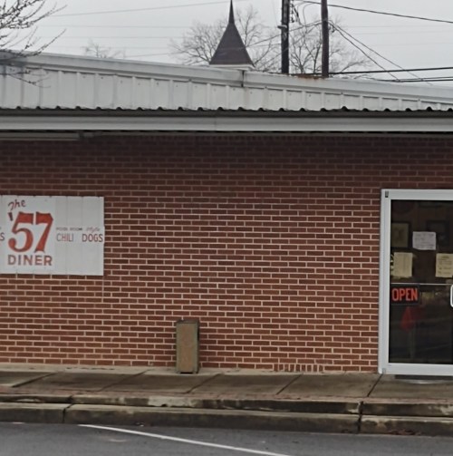 A brick diner with a sign reading "57 Diner" and "Chili Dogs," featuring an open door and a church steeple in the background.