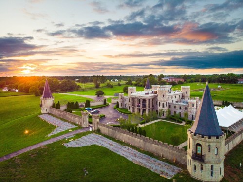 Aerial view of a castle with turrets, surrounded by green fields and a sunset in the background.
