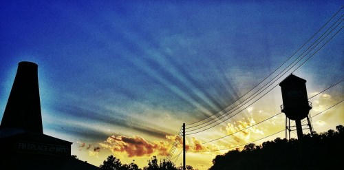 Silhouette of a water tower and buildings against a vibrant sunset with rays of light breaking through the clouds.