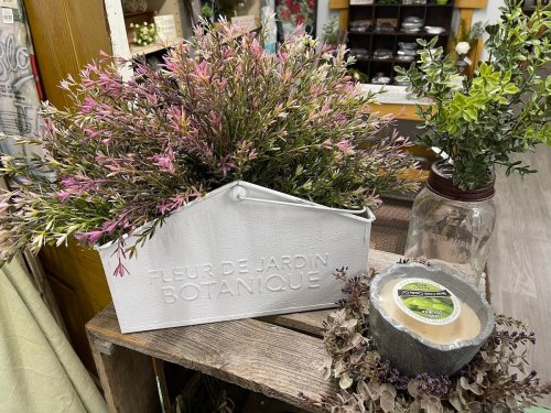 A white planter filled with pink and green flowers, next to a jar and a decorative wreath on a wooden display.