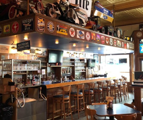 A cozy bar with wooden stools, shelves of drinks, and vintage signs, featuring a collection of memorabilia on the ceiling.
