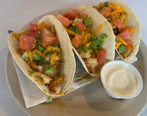 Three tacos filled with grilled chicken, lettuce, tomatoes, and shredded cheese, served with a side of creamy sauce.