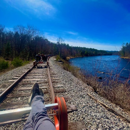 A person relaxes with their feet on a rail cart, overlooking a river and trees under a clear blue sky.
