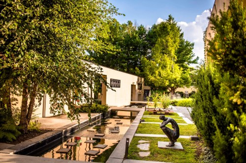 A serene garden with a reflecting pool, sculptures, and lush greenery under a clear blue sky.
