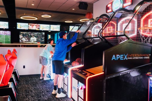 Two people playing basketball arcade games at Apex Entertainment, with screens showing sports in the background.