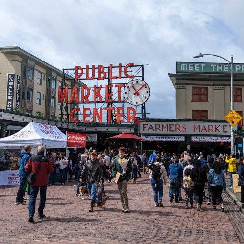 Crowd of people at a bustling public market with colorful signs and stalls under a cloudy sky.