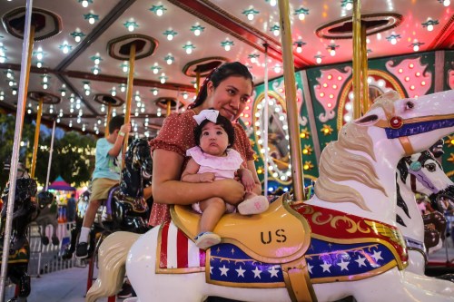 A mother holds her baby on a carousel horse, surrounded by colorful lights and festive decorations.