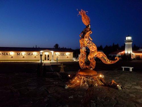 A whimsical mermaid statue stands in front of the National Mermaid Museum at dusk, with a lighthouse in the background.