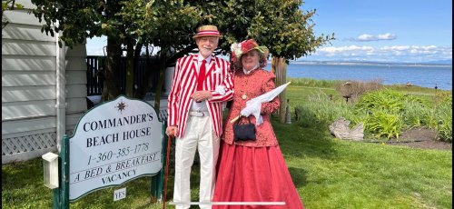 A man in a striped suit and a woman in a red dress pose in front of a beach house sign by the water.