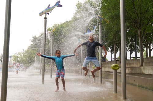 Two children play joyfully in a splash pad, jumping through water sprays under colorful fish decorations.