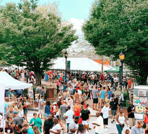 A bustling outdoor festival with crowds, tents, and trees, filled with people enjoying the event.