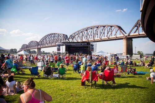 A crowd enjoys a music festival on a grassy field, with a large bridge and stage in the background under a clear sky.