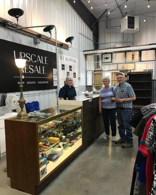 A couple stands at a resale shop counter, chatting with a volunteer amidst shelves of items for sale.