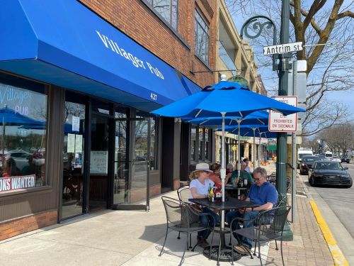 Outdoor seating at Villager Pub with blue umbrellas, patrons dining, and a street sign for Antrim St. on a sunny day.