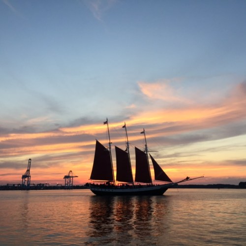 A tall ship with red sails glides across the water at sunset, with cranes silhouetted in the background.