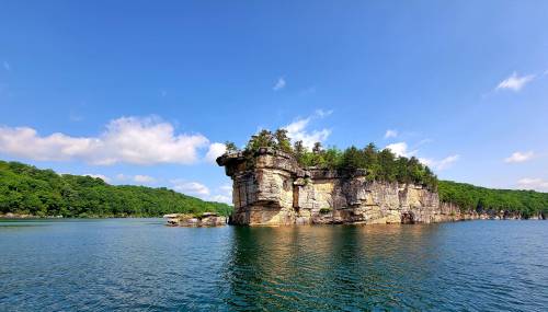 A rocky island with trees surrounded by calm blue water under a clear sky with a few clouds.