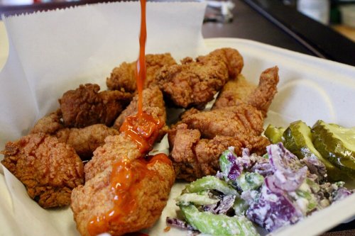 A serving of crispy fried chicken drizzled with sauce, accompanied by pickles and a creamy coleslaw.