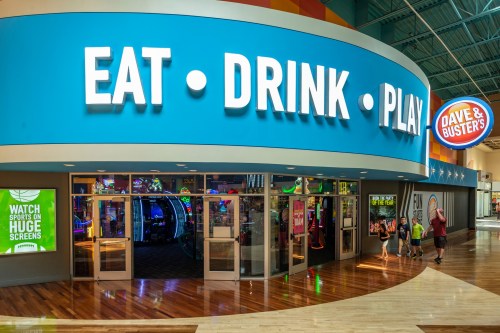 Entrance to Dave & Buster's featuring a vibrant sign that reads "Eat. Drink. Play." with arcade games visible inside.