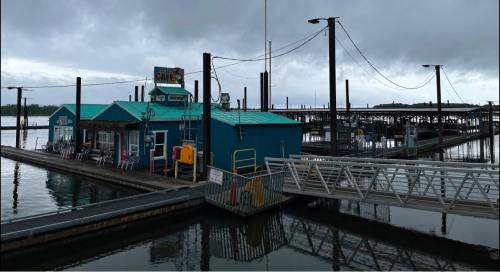 A blue café on a dock with a cloudy sky and empty boat slips in the background. Reflections in the water.