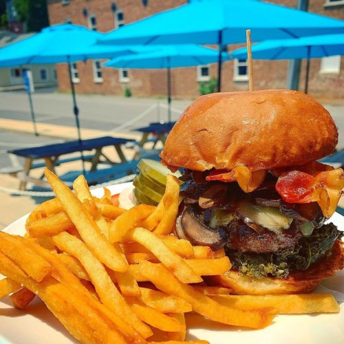 A juicy burger with toppings, served with crispy fries, set against a backdrop of outdoor seating and blue umbrellas.