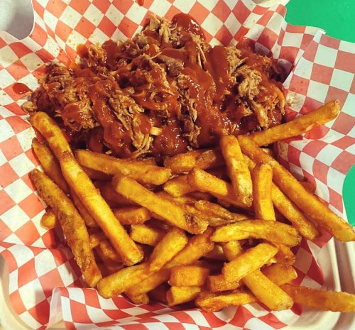 A basket of pulled pork topped with barbecue sauce, served with a side of golden French fries on a checkered paper liner.