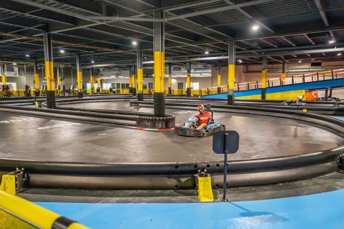 A go-kart track indoors with a racer navigating a curve, surrounded by safety barriers and bright yellow pillars.