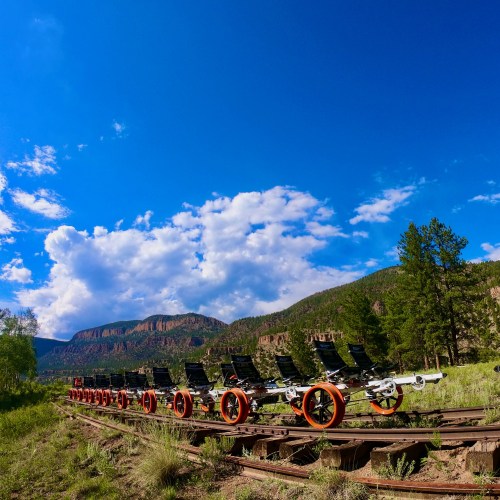A row of empty train cars on tracks, set against a backdrop of blue sky and green mountains.