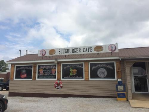 Exterior of Slugburger Cafe with a sign and windows displaying menu items, set against a cloudy sky.
