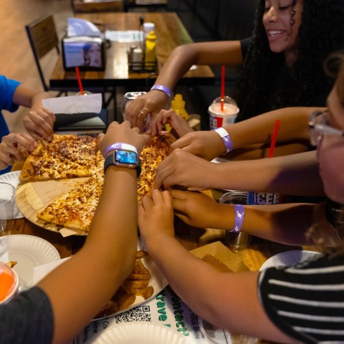 A group of hands reaching for a pizza on a table, with drinks and condiments in the background.