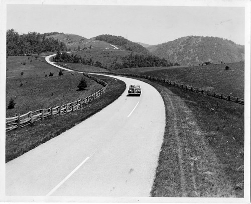 A vintage car drives along a winding road through a hilly landscape, bordered by a wooden fence.