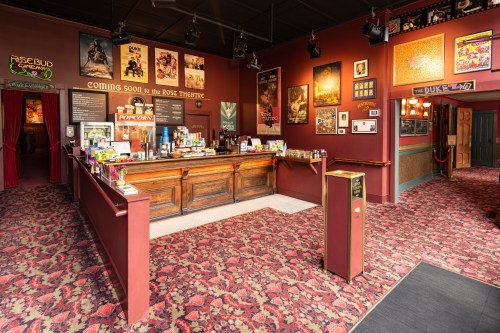 Interior of a vintage theater lobby with a bar, colorful posters on the walls, and patterned carpet.