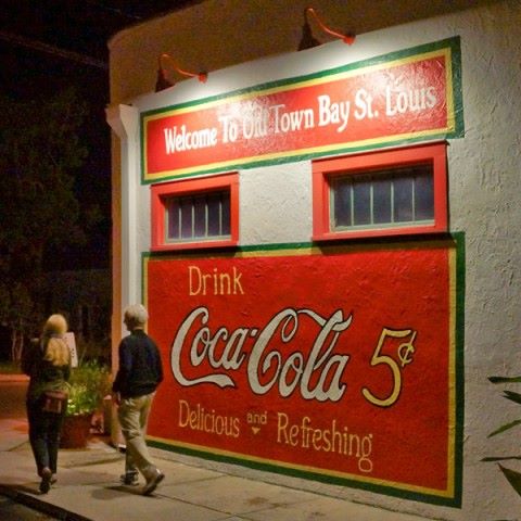 Two people walk past a colorful vintage Coca-Cola advertisement on a building in Old Town Bay St. Louis at night.