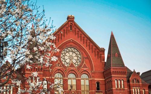 Historic red brick building with ornate architecture, framed by blooming white flowers against a clear blue sky.