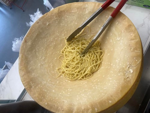 A large cheese wheel holds freshly cooked pasta, with tongs resting inside the bowl.