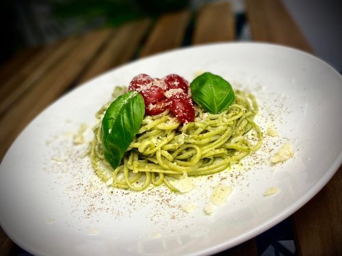 A plate of pasta with pesto, topped with cherry tomatoes, basil leaves, and grated cheese, on a wooden table.