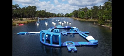 Aerial view of a blue inflatable water park on a lake, surrounded by trees and sailboats in the background.