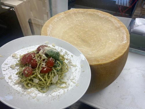 A plate of pasta with cherry tomatoes and basil next to a large wheel of cheese.