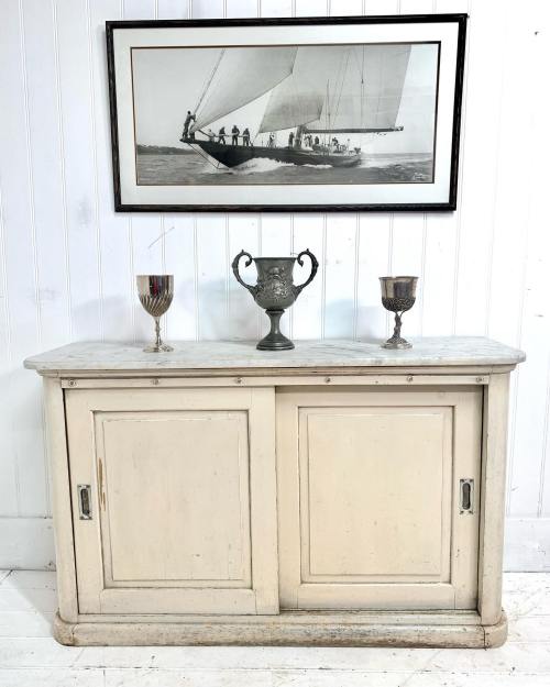 A white cabinet with a marble top, flanked by two silver cups, beneath a framed sailing artwork on a white wall.