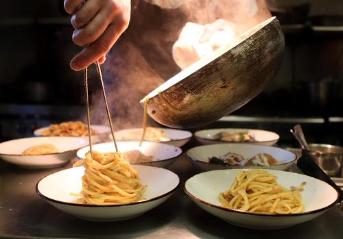 A chef twirls pasta onto plates in a steamy kitchen, showcasing a delicious dish being prepared.