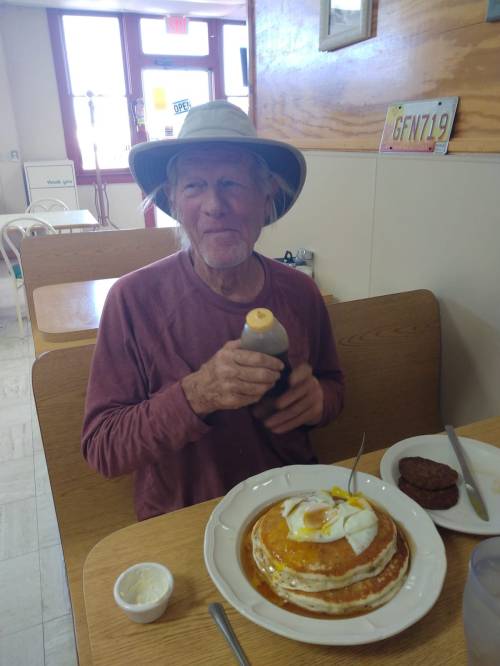 A smiling man in a hat holds a bottle of syrup next to a large plate of pancakes with an egg and sausage.