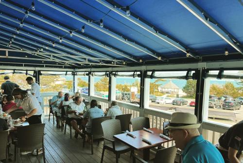 A lively restaurant patio with diners, blue awning, and a view of cars and trees outside.