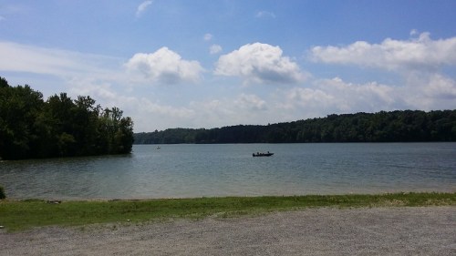 A serene lake surrounded by trees under a blue sky with fluffy clouds, and a small boat in the water.