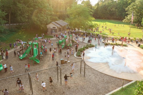 A busy playground with children playing on slides and a splash pad, surrounded by green trees and a sunny atmosphere.