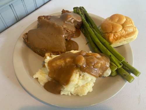 A plate with meatloaf topped with gravy, mashed potatoes with gravy, asparagus, and a dinner roll.