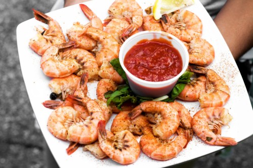 A plate of cooked shrimp garnished with lemon and served with a small cup of cocktail sauce.