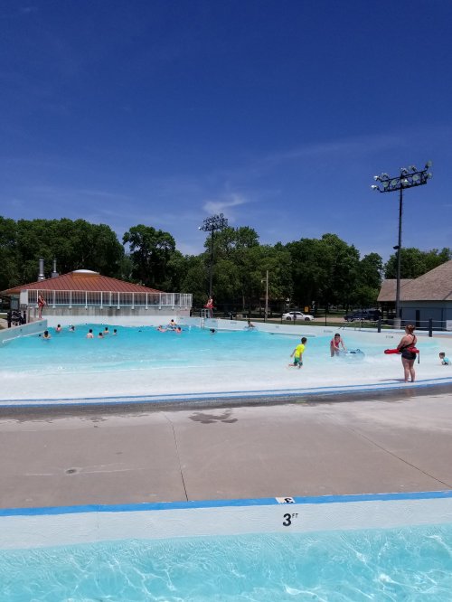A sunny day at a public pool with children swimming and playing, surrounded by trees and a concession area.