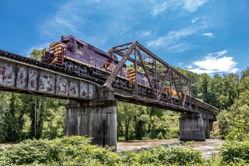 A purple train crosses a metal bridge over a river, surrounded by lush green trees and a blue sky.
