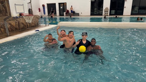 A group of six kids smiles and poses in a swimming pool, holding a yellow ball, enjoying their time together.