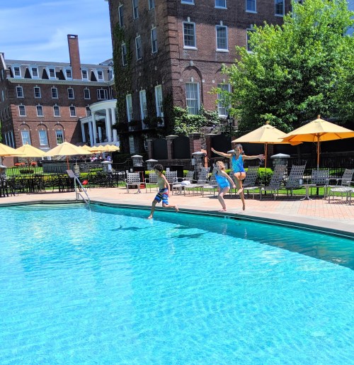 Three children joyfully leap into a bright blue swimming pool on a sunny day, surrounded by lounge chairs and umbrellas.